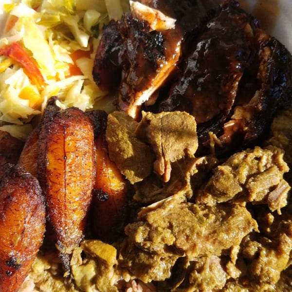A close-up of a Caribbean dish with jerk chicken, curried goat, fried plantains, and cabbage.