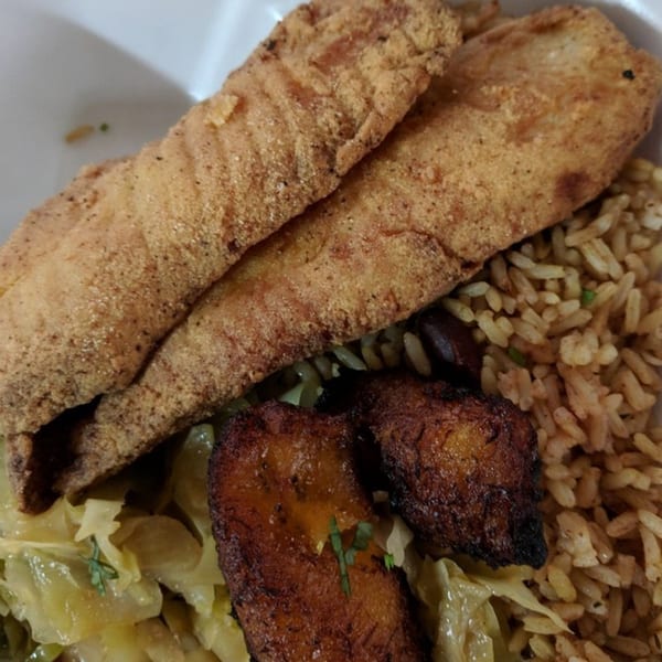 Fried fish fillets, rice and peas, stewed cabbage, and plantains in a white takeout container.